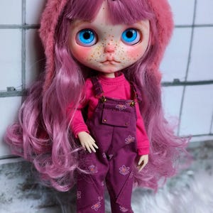 May include: A doll with long, pink hair and bright blue eyes, wearing a pink hooded hat with cat ears. The doll is dressed in a pink long-sleeved shirt and purple overalls with floral designs. The doll's face has freckles and is wearing purple shoes.