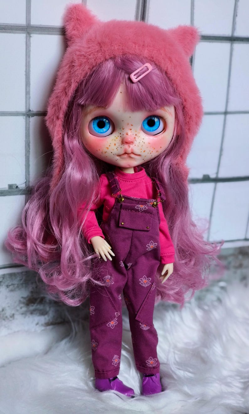 May include: A doll with long, pink hair and bright blue eyes, wearing a pink hooded hat with cat ears. The doll is dressed in a pink long-sleeved shirt and purple overalls with floral designs. The doll's face has freckles and is wearing purple shoes.