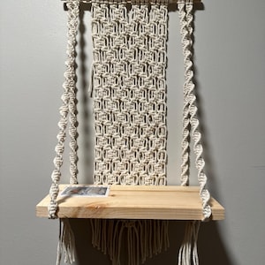 May include: A macrame wall shelf with a natural wood shelf and a white macrame design. The shelf is hanging from a wooden dowel rod.