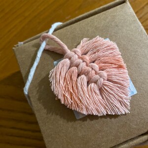 May include: A small, pink heart-shaped macrame wall hanging with a white string attached to the top.