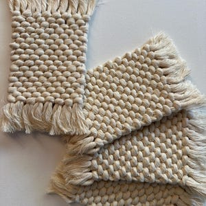 May include: Off-white woven coasters with a textured, knotted design and fringed edges. The coasters are rectangular and square-shaped, and are arranged on a white surface. The natural colour and texture suggest a handmade craft.