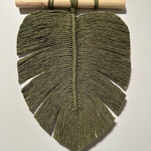 May include: Olive green macrame leaf wall hanging with a wooden dowel and hanging cord. The leaf design features a central stem and detailed fringe, creating a textured, natural aesthetic. This handcrafted piece adds a touch of bohemian style to any space.