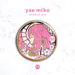 May include: A round enamel pin featuring a pink-haired anime character with gold accents. The pin has the text "yae miko enamel pin" in pink and gold. The character is set against a pink and gold background with floral details.
