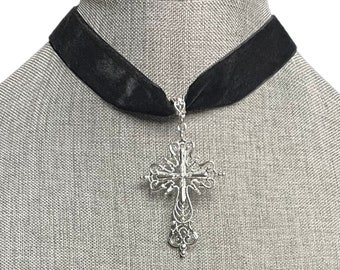 May include: A black velvet choker necklace with a silver-colored filigree cross pendant. The cross features intricate openwork and a detailed design. The necklace is displayed on a gray mannequin.
