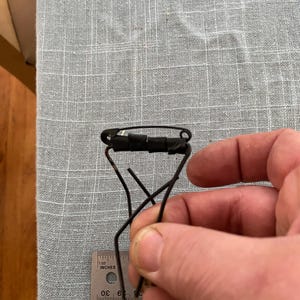 May include: A black metal eyelash curler is held by a hand. The curler is made of thin metal rods and has a black rubber pad. A ruler with inch measurements is in the image.