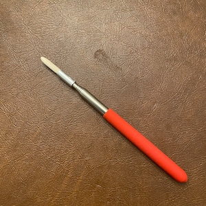 May include: A silver-toned tool with a red handle and a white, rectangular tip. The tool is designed for precision work and is set against a brown, textured background.