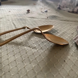 May include: Two antique gold-colored serving spoons with curved handles and oval bowls. The spoons are resting on a light green tablecloth with a diamond pattern. The spoons appear to be made of metal.