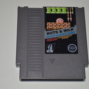 May include: A gray Nintendo Entertainment System game cartridge for the game "Nuts & Milk" from the Arcade Classics Series. The cartridge features a pixelated image of a pink character on a brick wall with a green background.