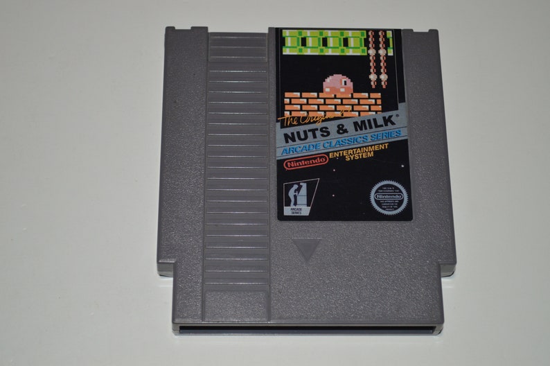May include: A grey Nintendo Entertainment System game cartridge for the game "Nuts & Milk" from the Arcade Classics Series. The cartridge features a pixelated image of a pink character on a brick wall with a green background.