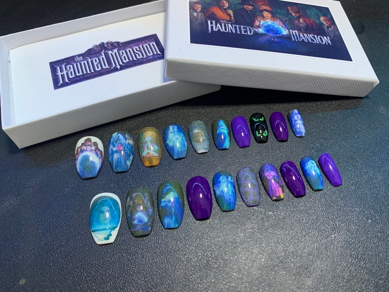 May include: A set of 14 coffin-shaped false nails with a Haunted Mansion theme. The nails feature images of characters and scenes from the Disney attraction, including the Ghost Host, Constance Hatchaway, and the mansion itself. Some nails are solid purple, while others have a clear base with the images printed on them.