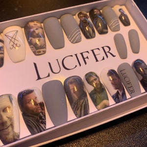 Peut inclure: Un ensemble d'ongles press-on gris avec des images d'un visage d'homme et le mot "LUCIFER" imprimé dessus.