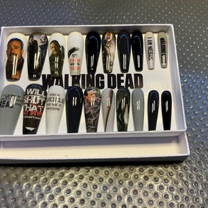 Op de afbeelding: Een set van 16 kunstnagels met een Walking Dead-thema. De nagels zijn zwart, grijs en wit met verschillende afbeeldingen en tekst uit de serie, waaronder "Will Shut That Down", "This is Lucille and she is awesome" en "I am Negan".
