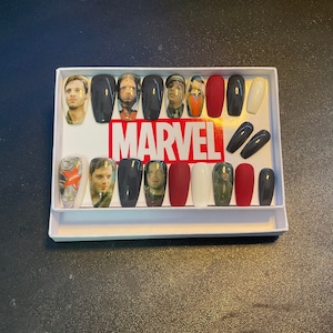 Puede incluir: Una caja blanca con un logotipo MARVEL negro y rojo. La caja contiene 14 uñas postizas con imágenes de personajes del Universo Cinematográfico de Marvel.