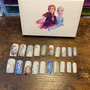 May include: Set of 12 white press-on nails with various Frozen designs, including Elsa, Anna, Olaf, and snowflakes. The nails have a light blue and silver glitter accent. The set comes in a box with the word "Frozen" printed on it.