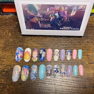 Puede incluir: Un conjunto de 14 uñas postizas ovaladas con imágenes de la película animada "El castillo ambulante". Las uñas son de varios colores, incluyendo rosa, azul y morado. Las imágenes en las uñas incluyen personajes de la película, como Howl, Sophie y Calcifer.