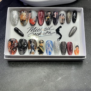 Könnte beinhalten: Ein Set von Kunstnägeln mit verschiedenen Designs, darunter Glitzer, Porträts und Bilder von Michael Jackson. Die Nägel sind in einer weißen Box angeordnet, auf der der Name "Michael Jackson" steht. Die Nägel sind oval geformt und in verschiedenen Farben erhältlich.