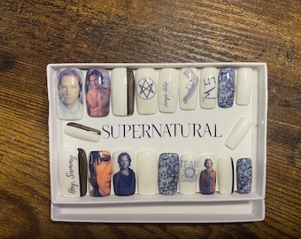 May include: A set of white press-on nails with various designs featuring characters and symbols from the TV show Supernatural. The designs include the show's logo, the words "Team Free Will", "My shoe", and "Hey Sammy", and images of the characters Dean Winchester and Sam Winchester.