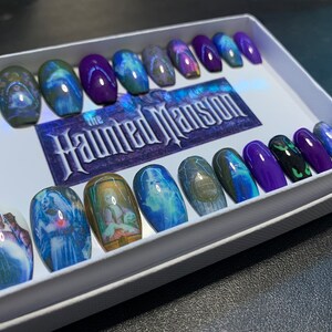 May include: A set of 18 coffin-shaped press-on nails with a Haunted Mansion theme. The nails feature images of ghosts, the Haunted Mansion sign, and other details from the Disney attraction. The nails are in a white box with the words "The Haunted Mansion" printed on the lid.