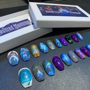 May include: A set of 18 coffin-shaped press-on nails with a purple base and images of ghosts from the Haunted Mansion attraction. The nails are arranged in two rows on a dark surface. The box for the nails is in the background and has the words "The Haunted Mansion" printed on it.