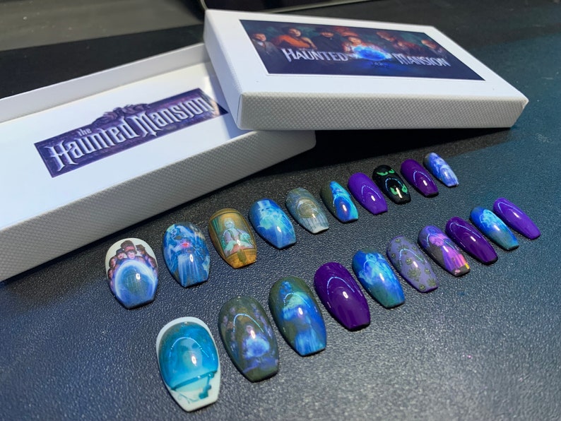 May include: A set of 18 coffin-shaped press-on nails with a purple base and images of ghosts from the Haunted Mansion attraction. The nails are arranged in two rows on a dark surface. The box for the nails is in the background and has the words "The Haunted Mansion" printed on it.