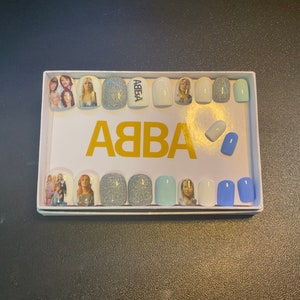 Könnte beinhalten: Ein Set mit 12 falschen Nägeln mit weißem Hintergrund und blauen und silbernen Akzenten. Die Nägel zeigen ein Bild der Band ABBA und das Wort "ABBA" in goldenen Buchstaben.