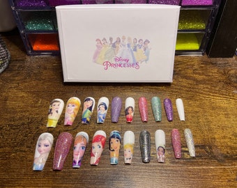 Könnte beinhalten: Set mit 18 falschen Nägeln im Disney-Prinzessinnen-Design. Die Nägel sind weiß mit verschiedenen Glitzer- und Prinzessinnen-Charakterdesigns. Die Schachtel im Hintergrund trägt die Aufschrift "Disney Princesses".