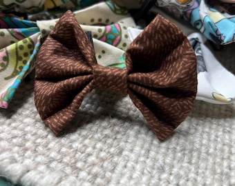 May include: A brown fabric bow tie with a textured herringbone pattern, centered on a neutral-colored woven surface. Other colorful bow ties with various patterns are visible in the background, suggesting a collection of accessories.