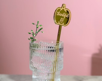 Op de afbeelding: Gouden drinkroerder in de vorm van een pompoen met de tekst "thankful AF" op een roze achtergrond. De roerder zit in een helder glas met water en een takje groen.