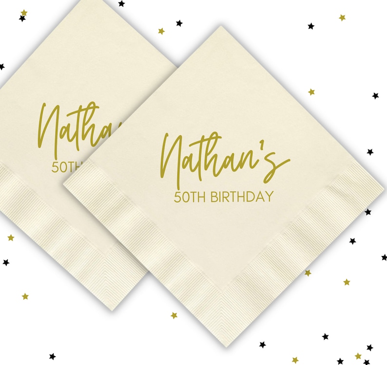 Puede incluir: Dos servilletas de papel blancas con letras doradas. La servilleta de arriba dice "Nathan's 50th Birthday".