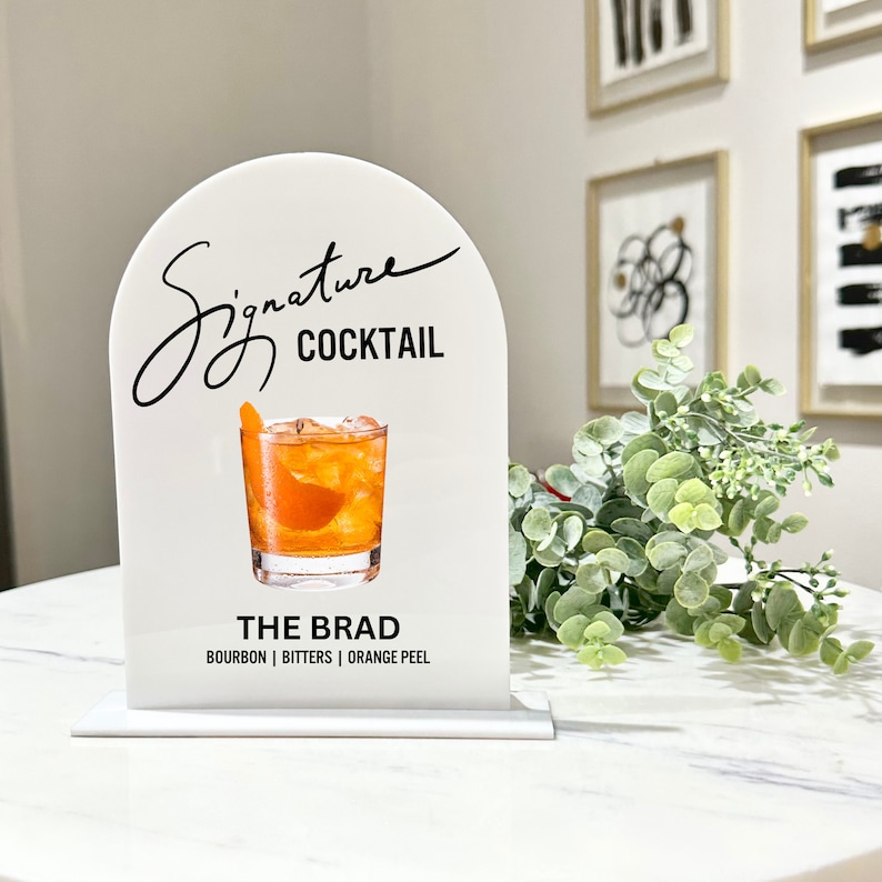May include: White acrylic sign with an arch shape at the top. The sign says "Signature Cocktail" in black cursive writing. Below the text is a picture of an orange cocktail in a rocks glass. The text "The Brad" is below the picture, and the ingredients "Bourbon | Bitters | Orange Peel" are listed below that.
