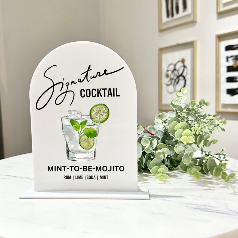 May include: White sign with a black arch and the text "Signature Cocktail" in black script. A drawing of a glass of mojito with lime and mint is below the text. The text "MINT-TO-BE-MOJITO" is below the drawing, and the ingredients "RUM | LIME | SODA | MINT" are listed below that.
