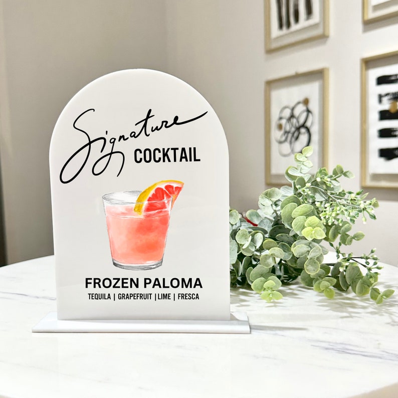 May include: White acrylic sign with a rounded top, featuring a hand-drawn illustration of a pink cocktail with a grapefruit slice. The text "Signature Cocktail" is written in black cursive script above the illustration. Below the illustration, the text "Frozen Paloma" is written in black, followed by the ingredients "Tequila | Grapefruit | Lime | Fresca".