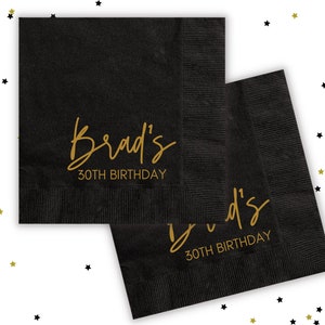 Puede incluir: Dos servilletas de papel negras con letras doradas. El texto en las servilletas dice "Brad's 30th Birthday".