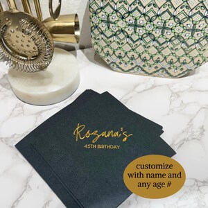 Puede incluir: Servilletas negras con letras doradas que dicen "Rozanna's 45th Birthday". Las servilletas est&aacute;n apiladas sobre una superficie de m&aacute;rmol blanco. Una pegatina ovalada dorada dice "personalizar con nombre y cualquier edad #".