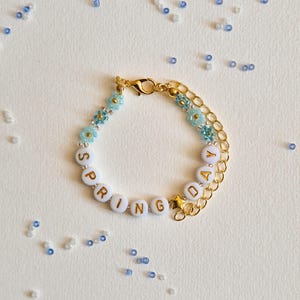 May include: A gold chain bracelet spelling "SPRING DAY" in white letter beads. It features light blue flower and bead details, and a gold star charm. Blue and white beads are scattered around the bracelet. The bracelet is approximately 17 cm in length.