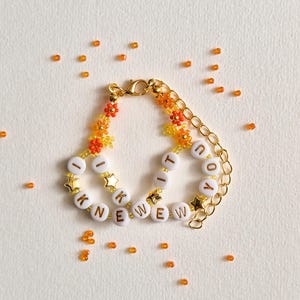 Puede incluir: Una pulsera de cadena dorada con cuentas de letras blancas que forman la frase I KNEW YOU, intercaladas con peque&ntilde;as cuentas en forma de flor naranja y amarilla y dijes de estrellas doradas. Incluye un cierre de mosquet&oacute;n dorado.