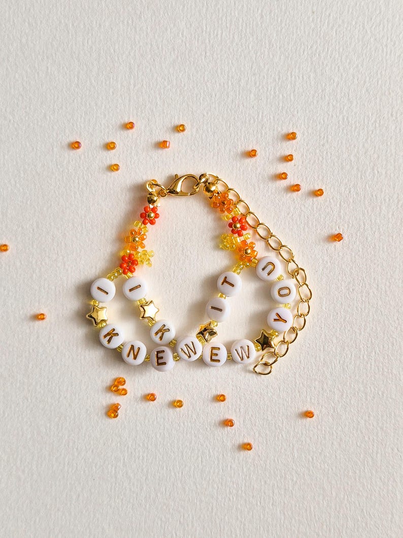 Puede incluir: Una pulsera de cadena dorada con cuentas de letras blancas que forman la frase I KNEW YOU, intercaladas con peque&ntilde;as cuentas en forma de flor naranja y amarilla y dijes de estrellas doradas. Incluye un cierre de mosquet&oacute;n dorado.