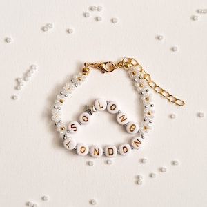 May include: A white beaded bracelet with gold lettering spelling "SO LONG LONDON". The bracelet has a gold clasp and is surrounded by white beads.
