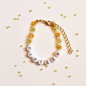 May include: A gold beaded bracelet with yellow and white flower beads spelling out the word "FIFTEEN".
