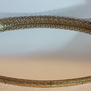 May include: A gold-toned, oval vanity tray with a mirrored surface. The tray features an ornate, openwork design around the edges, adding a touch of vintage elegance. The tray is empty and ready for use.