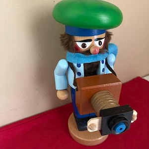May include: A wooden nutcracker figure wearing a bright green beret, a light blue scarf, and a light blue jacket with dark blue polka dots. The figure has brown hair and holds a brown box-shaped camera with a lens extending forward.