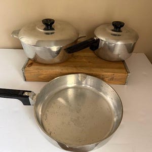 May include: A set of vintage aluminum cookware, including a large pot with a lid, a smaller pot with a lid, and a frying pan. The pots have black handles and knobs. The cookware is displayed on a white surface.