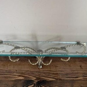 May include: A rectangular glass shelf with decorative gold-colored metal supports. The supports feature a rope-like design and curved hooks. The shelf is mounted on a wooden surface.