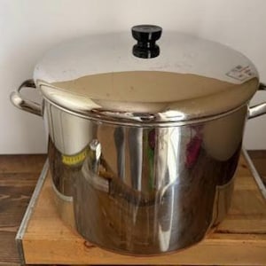 May include: A large, shiny stainless steel pot with a lid and two side handles. The lid has a black knob. The pot sits on a wooden surface. Kitchen cookware.