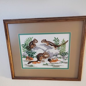 May include: Framed cross-stitch artwork featuring four chipmunks in a natural setting. The chipmunks are brown and white, with a detailed depiction of rocks, ferns, and autumn leaves. The artwork is framed in a brown wooden frame.
