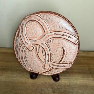 May include: A circular ceramic plate featuring a textured, light copper-colored finish with a raised, embossed design of three interlocking horseshoes. The plate is displayed on a small, dark brown stand, showcasing a rustic and weathered appearance.