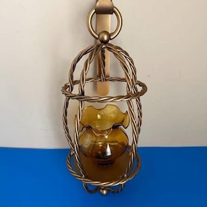 May include: A wall-mounted candle holder featuring a gold-toned metal cage with a twisted rope design. The cage is suspended from a circular ring and holds a small, amber-colored glass vessel with a ruffled, scalloped rim at the center.