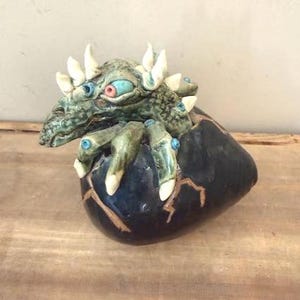 May include: A ceramic sculpture of a creature emerging from a cracked egg. The creature has a green, textured body with white spikes and blue and pink eyes. The egg is dark blue with gold-colored cracks.