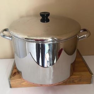 May include: A large, shiny stainless steel pot with a lid and two side handles. The lid has a black knob. The pot sits on a wooden trivet. The pot reflects the surrounding environment.