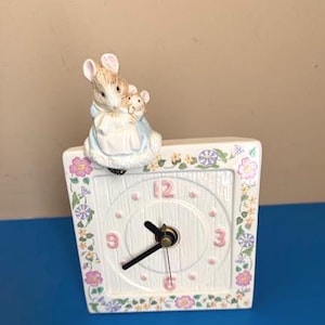 May include: A square ceramic clock featuring a white face with embossed vertical lines, pink numbers 3, 6, 9, and 12, and black hands. The border is decorated with colorful floral patterns. A ceramic mouse figure in a blue coat holds a smaller mouse.